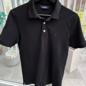 Svart pikétröja  - Snygg svart pikétröja från Jack and Jones med klassisk krage och knappar framtill. Perfekt för en stilren look. Passar bra till både jeans och chinos.