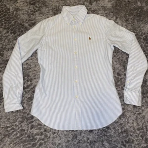 Randig ralph lauren skjorta - Snygg randig ralph lauren skjorta i blått och vitt med en broderad logga på bröstet. Skjortan är i costum fit och har långa ärmar och knappar framtill. Passar i S-XS