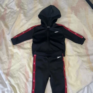 Svart hoodie från Reebok med röda detaljer - Säljer en svart hoodie från Reebok med röda detaljer längs ärmarna. Tröjan har en dragkedja framtill och en huva. Perfekt för en sportig look!
