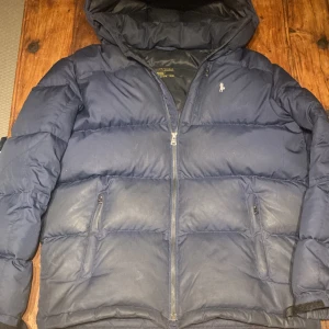 Mörkblå dunjacka från Ralph Lauren - Säljer en snygg mörkblå dunjacka från Ralph Lauren. Jackan har en dragkedja framtill och en huva. Den är i bra skick dock är den smått fläckig och på en av bilderna ser man att de även finns ytterst små hål i jackan. Den sitter som en liten S eller stor XS. 