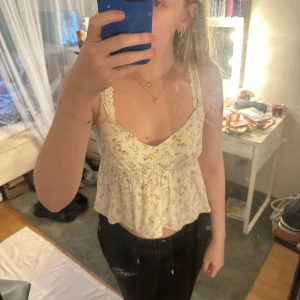 Blommig vit linnetopp - Säljer en söt vit linnetopp med blommigt mönster i gult och grönt. Toppen har spetsdetaljer vid axelbanden och en v-ringad design. Perfekt för varma sommardagar!