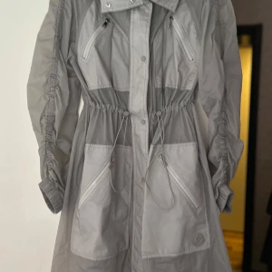 Grå regnjacka från Moncler - Säljer en stilren grå regnjacka från Moncler med justerbar midja och dragkedjor. Jackan har långa ärmar och tryckknappar framtill. Perfekt för regniga dagar och håller dig torr med stil.