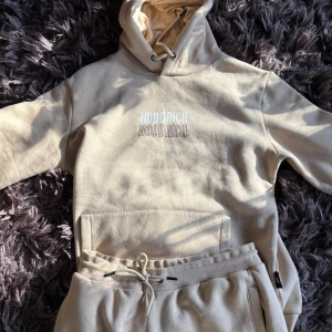  Hoodrich set - Säljer en beige hoodie från Hoodrich med tryck på bröstet. Den har en stor ficka framtill och justerbar huva med dragsko. Perfekt för en avslappnad stil.