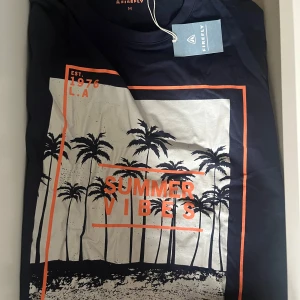 Svart t-shirt med tryck från Firefly - Snygg svart t-shirt från Firefly med ett coolt tryck av palmer och texten 'Summer Vibes' i orange. Perfekt för en avslappnad stil med sommarkänsla. T-shirten har korta ärmar och en normal passform.