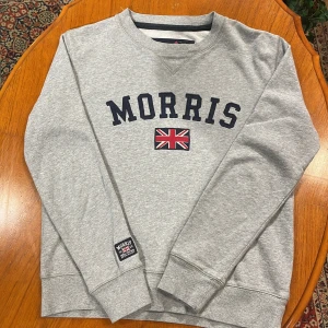 Grå sweatshirt från Morris - Säljer en stilren grå sweatshirt från Morris med broderad logga och brittisk flagga på bröstet. Tröjan har långa ärmar och ribbade muddar vid ärmslut och nederkant. Perfekt för en avslappnad stil.
