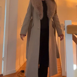 Beige kappa med pälsdetalj - Elegant beige kappa med en lyxig pälsdetalj runt kragen. Som är avtagbar med hjälp av knappar💕perfekt nu inför hösten🍂🍁