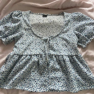 Blommig topp från Gina Tricot - Säljer en söt blommig topp från Gina Tricot i storlek M. Toppen har korta puffärmar och är öppen framtill med 2 knytningar. Den är i en ljusblå nyans med små blommor i mörkare blått och vitt. Perfekt för vår och sommar!🌷HELT NY 