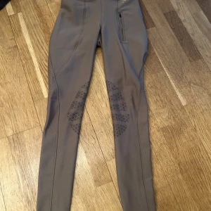 Bruna ridbyxor från Equithème - Snygga bruna ridbyxor från Equithème med hög midja och tight passform. Byxorna har en praktisk dragkedja fram och dekorativa mönster på insidan av benen för extra grepp. Perfekta för ridning och designade för komfort och stil.