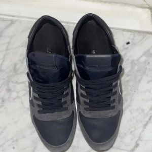 Grå och svarta sneakers från Philippe Model - Snygga sneakers från Philippe Model i en stilren kombination av grått och svart. Skorna har en klassisk design med snörning och en vit sula som ger en fräsch kontrast. Perfekta för en avslappnad och trendig look.