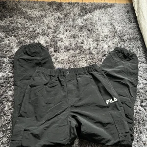 Svarta byxor från FILA - Säljer ett par svarta byxor från FILA med elastisk midja och avslappnad passform. Perfekta för en bekväm stil. Byxorna har en enkel design med FILA-loggan på sidan.