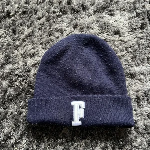 Mörkblå stickad mössa med broderad bokstav - Snygg mörkblå stickad mössa med en broderad bokstav 'F' framtill. Perfekt för att hålla värmen under kyliga dagar. Enkel och stilren design som passar till det mesta.