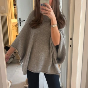 Grå stickad poncho - Säljer en mysig grå stickad poncho med vida ärmar. Perfekt för kyliga dagar och ger en avslappnad look. Passar bra till jeans och kjol.