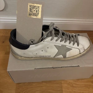 Vit och grå sneakers från Golden Goose - Snygga vita sneakers från Golden Goose med grå stjärndetalj på sidan och svarta hälpartier. Skorna har grå snörning och en klassisk design med en bekväm gummisula. Perfekta för en stilren look. Box till skorna samt påse ingår. Skorna är andvända fåtal gånger. Skorna säljs i befintligt skick