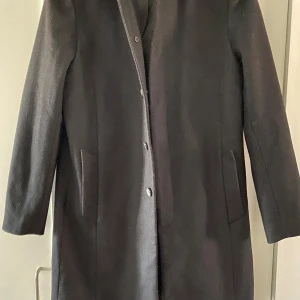 Svart kappa från Nikolaj - Elegant svart kappa från Nikolaj med knappar framtill och diskreta fickor. Insidan har en stilren vinröd detalj som ger en lyxig känsla. Perfekt för kyligare dagar.