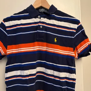 Randig pikétröja från Ralph Lauren - Snygg pikétröja från Ralph Lauren med ränder i marinblått, vitt och orange. Tröjan har en klassisk krage och knappar framtill. Perfekt för en stilren look.