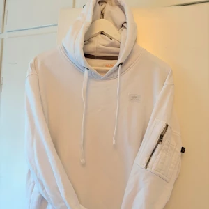 Vit hoodie från Alpha Industries - Säljer en stilren vit hoodie från Alpha Industries. Den har en praktisk ficka med dragkedja på ärmen och en klassisk känguruficka framtill. Hoodien har en justerbar huva med dragsko och en liten logotyp på bröstet.