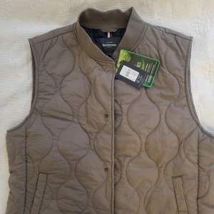 Beige quiltad väst från Dressmann - Säljer en beige quiltad väst från Dressmann. Västen har en stilren design med knappar framtill och en ribbad krage. Perfekt för att hålla värmen under kyligare dagar.