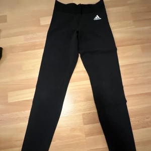 Svarta leggings från Adidas - Snygga svarta leggings från Adidas med vit logga på framsidan och stora vita tryck på benen. Perfekta för träning eller en avslappnad dag. De har en tight passform och är tillverkade i ett stretchigt material för bästa komfort.