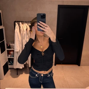 Svart långärmad korsettopp - Snygg svart korsettopp med långa ärmar och en liten knytdetalj framtill. Toppen har en tight passform och är perfekt för en kväll ute. Den har en stilren design som framhäver figuren.