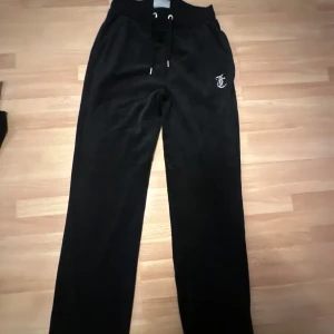 Svarta mjukisbyxor från Juicy Couture - Snygga svarta mjukisbyxor från Juicy Couture med glittrande text på baksidan. Byxorna har en bekväm passform med snörning i midjan och ett litet emblem framtill. Perfekta för en avslappnad stil.