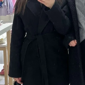 Svart kappa med bälte - Snygg svart kappa med bälte i midjan. Perfekt för kyligare dagar med sin stilrena design och långa ärmar. Ett måste i garderoben för en klassisk look.