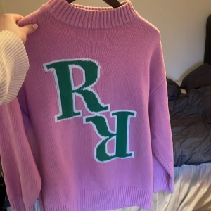 Lila tröja med gröna bokstäver - Säljer en cool lila tröja med stora gröna bokstäver på framsidan. Tröjan har en ribbad krage och är perfekt för en avslappnad stil. Passar bra till jeans eller kjol för en trendig look.