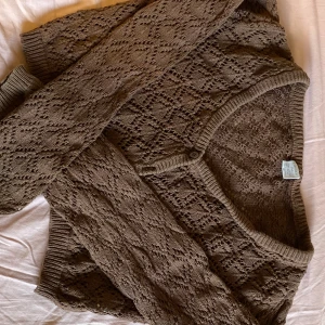 Brun stickad kofta från Lindex - Säljer en brun stickad kofta från Lindex med ett fint hålmönster. Koftan har långa ärmar och knappar framtill. Perfekt för att hålla sig varm och stilren under kyligare dagar.