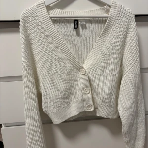 Vit stickad kofta från H&M Divided - Säljer en vit stickad kofta från H&M Divided. Koftan har en v-ringad design med stora knappar framtill. Perfekt för en stilren och mysig look. Säljer då den inte riktigt är min stil/ej används. 