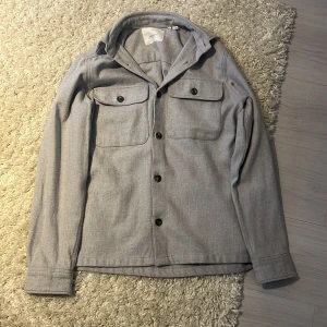 Grå overshirt från Jack & Jones - Säljer en stilren grå overshirt från Jack & Jones. Den har långa ärmar och knappar framtill samt två bröstfickor med knappar. Det är storlek xs men jag tycker det är väldigt stor i storleken. Priset är inte hugget i en sten.