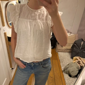 Vit blus med spetsdetaljer - Säljer en söt vit blus med spetsdetaljer och volanger på ärmarna. Blusen har en luftig passform och ett diskret randigt mönster på övre delen. Perfekt för en somrig look!