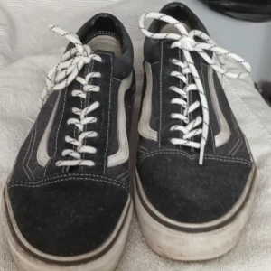 Svarta sneakers från Vans - Säljer ett par klassiska svarta Vans sneakers med vita detaljer och snörning. Skorna har en vit sula och den ikoniska vågformade sidodetaljen. Perfekta för en avslappnad stil. Det biliga priset är pågrund av att de är väldigt smutsiga och använda men inte trasiga.