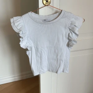 Vit ribbad topp från Zara - Säljer en söt vit ribbad topp från Zara med volangdetaljer på ärmarna. Toppen har en rund halsringning och är perfekt för en stilren look. Passar bra till både jeans och kjol. Perfekt för vår och sommar!