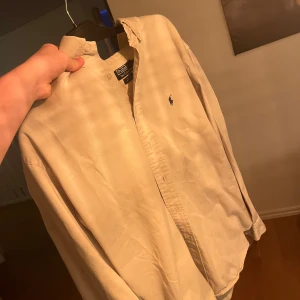 Beige skjorta från Ralph Lauren - Snygg beige skjorta från Ralph Lauren med klassisk krage och knappar framtill. Den har en broderad logga på bröstet och långa ärmar. Perfekt för en stilren look.