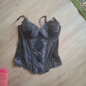 Svart korsett med spetsdetaljer - Elegant svart korsett med vackra spetsdetaljer och justerbara axelband. Den har en smickrande passform med genomskinliga partier och ett mönster som ger en lyxig känsla.