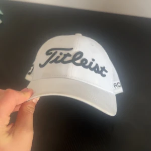 Vit keps från Titleist  - Snygg vit keps från Titleist med broderad logga framtill och texten 'PRO V1' på sidan. Baksidan har ett nätmaterial för bättre ventilation och justerbar rem för perfekt passform.