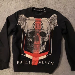 Svart Philip plein tröja  - Säljer en svart tröja från Philipp Plein med ett stort dödskalletryck och vingar på framsidan. Tröjan har långa ärmar och rund halsringning. Perfekt för en edgy look!