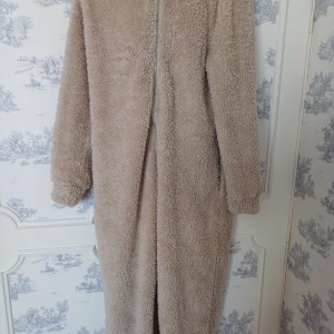 Beige one piece från ONLY - Mysig beige teddy one piece från ONLY med dragkedja framtill.  har en lång och bekväm passform med huva, perfekt för kyliga dagar. Den fluffiga texturen ger en trendig look.