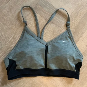 Grå sport-bh från Nike - Säljer en grå sport-bh från Nike med svarta detaljer och justerbara axelband. Den har en v-ringad design och är perfekt för träning. Nike-loggan är tryckt i vitt på framsidan.
