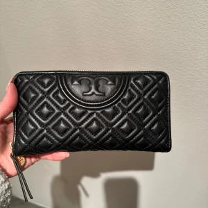 Svart plånbok från Tory Burch  - Snygg svart quiltad plånbok med dragkedja och elegant mönster. Perfekt för att hålla ordning på kort och kontanter. Den har en stilren design med guldfärgade detaljer.
