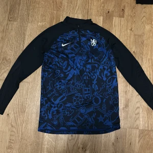  Chelsea träningströja  - Säljer en blå och svart långärmad Chelsea tröja från nike med ett unikt mönster och dragkedja vid halsen. Tröjan har en slim fit passform. 