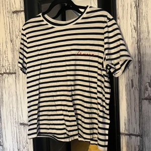 Randig t-shirt med broderi - Säljer en stilren svartvit randig t-shirt med ett diskret rött broderi på bröstet. Perfekt för en avslappnad look. T-shirten har korta ärmar och en klassisk rund halsringning.