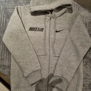 Grå hoodie från Nike (barn) - Säljer en grå hoodie från Nike med dragkedja och huva. Den har långa ärmar och ribbade muddar. Perfekt för en avslappnad stil.