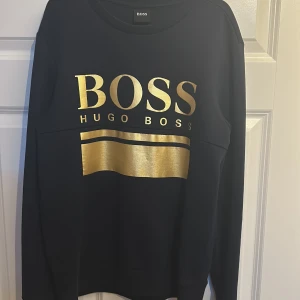 Svart tröja från Hugo Boss - Säljer en stilren svart tröja från Hugo Boss med guldtryck på bröstet. Tröjan har långa ärmar och en rund halsringning. Perfekt för en trendig look.