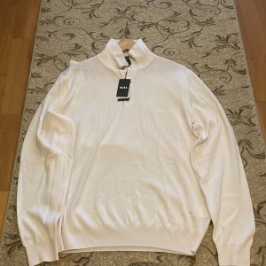 Helt ny tröja från Boss för 1100 kr, originalpriset 1800 kr - Säljer en stilren vit tröja från Boss med hög krage och diskret logga på bröstet. Perfekt för en elegant och avslappnad look. Tröjan har långa ärmar och är tillverkad i ett mjukt material.