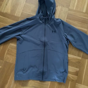 Grå vindjacka från Under Armour - Snygg grå vindjacka från Under Armour med huva och hel dragkedja framtill. Jackan har två sidofickor och en stilren logga på bröstet. Perfekt för blåsiga dagar och sportiga outfits. Lätt och smidig design som funkar året runt.