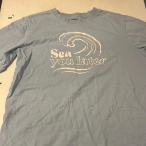 Ljusblå t-shirt med vågtryck - Ljusblå t-shirt med vit text och vågmotiv på bröstet där det står 'Sea you later'. Klassisk rund hals och korta ärmar. Skön och avslappnad passform, perfekt för chill dagar. Materialet känns mjukt och lätt. Dock har varan två fläckar som ni kan se på bilden.