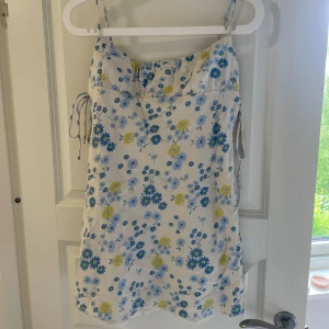 Vit blommig kortklänning med knyt - Supersöt vit kortklänning med blått och gult blommönster. Smala axelband, knyt i sidorna och lätt figursydd passform. Perfekt för varma dagar och har en fräsch, somrig vibe. Materialet känns lätt och luftigt.