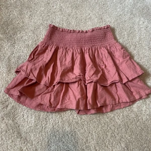 Rosa volangkjol med smock - Supersöt kort kjol i dammigt rosa med bred smock i midjan och dubbla volanger. Kjolen är luftig och har en flowy känsla, perfekt för varma dagar. Tillverkad i mjuk bomull som känns skön mot huden. Enkel att matcha med både crop tops och stickade tröjor.