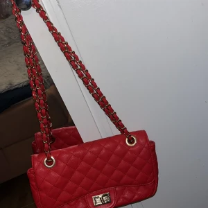 Röd quiltad axelväska med kedjerem - Säljer en snygg röd axelväska med quiltat mönster och guldfärgade detaljer. Väskan har en kedjerem flätad med rött material och ett klassiskt vridlås framtill. Insidan är fodrad i grått tyg och har både fack och dragkedja för småsaker.