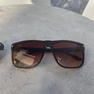 Svarta solglasögon med bruna glas - Snygga solglasögon med svart, fyrkantig båge och bruna tonade glas. Klassisk modell som ger cool vibe och passar perfekt till sommardagar. Bågarna är i plast och har små silvriga detaljer vid hörnen. Helt nya aldrig använda. 